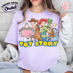 Puede incluir: Una camiseta de color lila claro con las palabras "TOY STORY" en letras de bloque amarillas y azules. La camiseta presenta un gr&aacute;fico colorido de personajes de la pel&iacute;cula animada, incluyendo a Woody, Buzz Lightyear, y otros. La camiseta tiene el logo "Comfort Colors" y "Orchid".