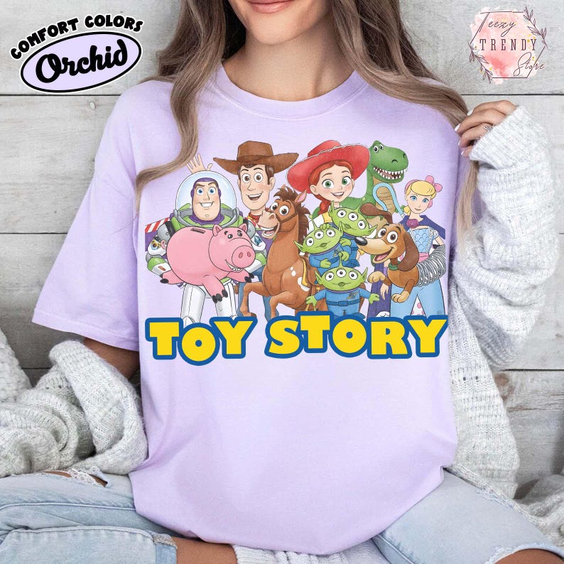 Puede incluir: Una camiseta de color lila claro con las palabras "TOY STORY" en letras de bloque amarillas y azules. La camiseta presenta un gr&aacute;fico colorido de personajes de la pel&iacute;cula animada, incluyendo a Woody, Buzz Lightyear, y otros. La camiseta tiene el logo "Comfort Colors" y "Orchid".