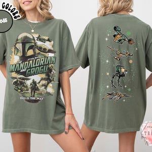 K&ouml;nnte beinhalten: Ein moosgr&uuml;nes T-Shirt mit einer Grafik auf der Vorderseite von The Mandalorian und Grogu sowie dem Text "The Mandalorian and Grogu" und "This is the way". Die R&uuml;ckseite zeigt eine vertikale Anordnung von vier Star Wars Raumfahrzeugen.