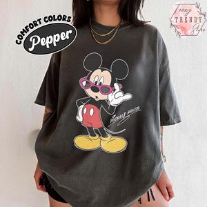 May include: A dark gray t-shirt featuring a graphic of Mickey Mouse wearing pink sunglasses and red shorts. The text "Mickey Mouse" is written in a cursive font. The shirt has the text "COMFORT COLORS Pepper" in a black oval.
