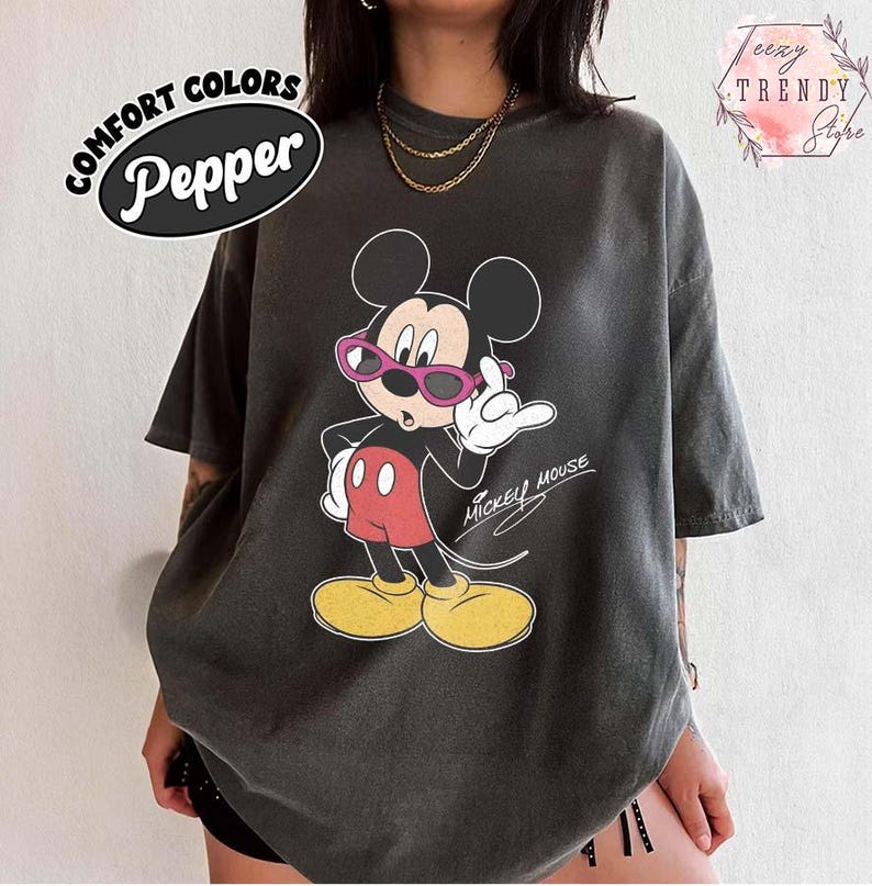 May include: A dark gray t-shirt featuring a graphic of Mickey Mouse wearing pink sunglasses and red shorts. The text "Mickey Mouse" is written in a cursive font. The shirt has the text "COMFORT COLORS Pepper" in a black oval.