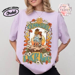 May include: A light purple t-shirt featuring a graphic print of two figures in cowboy hats standing together. The text COWGIRL LIKE ME is printed in large teal letters. The design includes orange flames, white and orange flowers, and a sun.