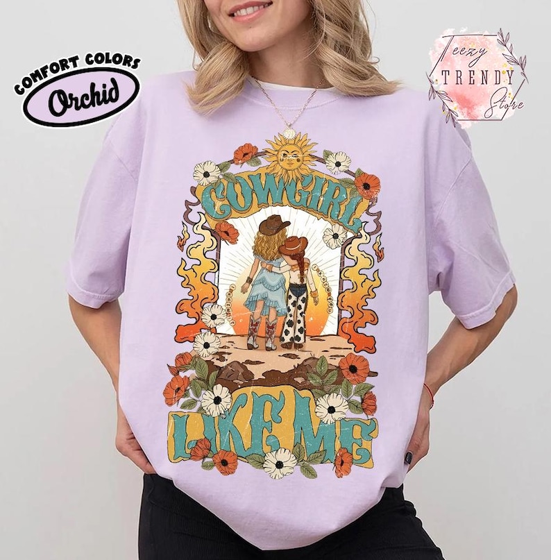 May include: A light purple t-shirt featuring a graphic print of two figures in cowboy hats standing together. The text COWGIRL LIKE ME is printed in large teal letters. The design includes orange flames, white and orange flowers, and a sun.