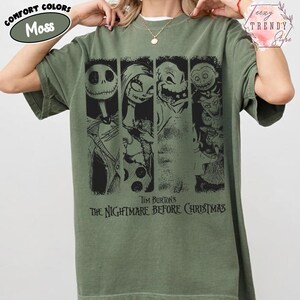 May include: A moss green short-sleeve t-shirt featuring a black graphic print of characters from Tim Burton's The Nightmare Before Christmas. The design includes four vertical panels showing Jack Skellington, Sally, Oogie Boogie, and Lock, Shock, and Barrel.