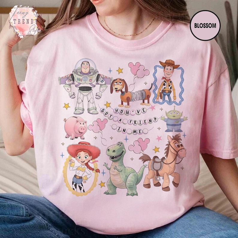 May include: Pink t-shirt featuring colorful cartoon characters from a popular animated movie. The design includes a space ranger, a cowboy, a dinosaur, a pig, a horse, and a green alien. The text "YOU'VE GOT A FRIEND IN ME" is also included.