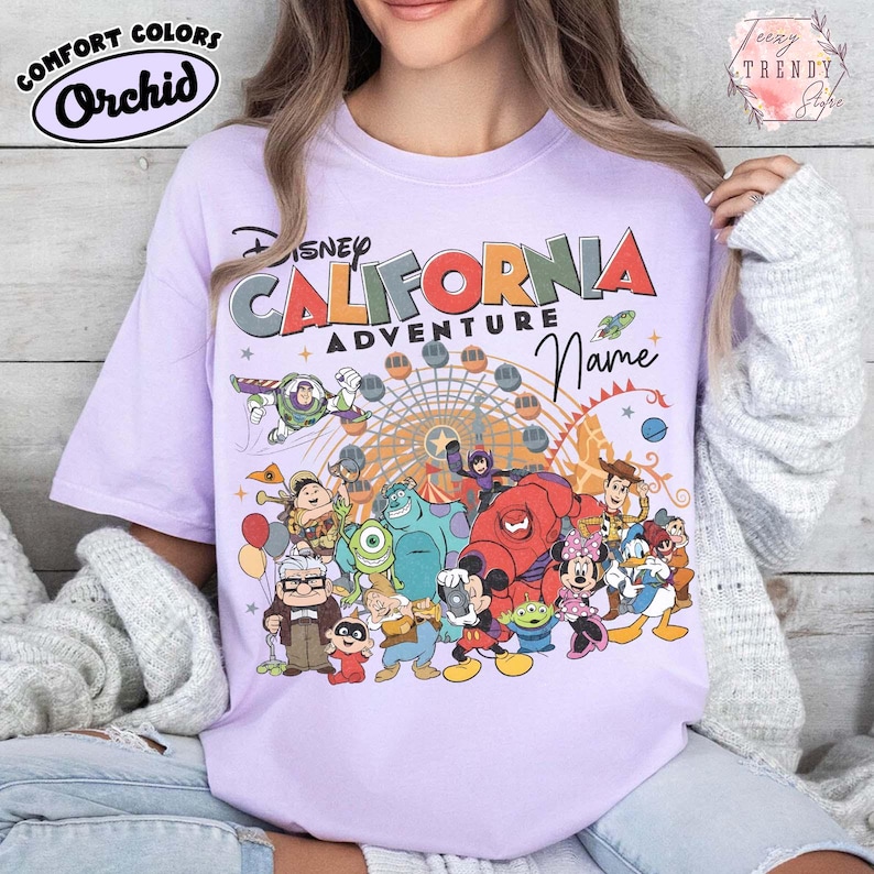 以下が含まれることがあります： ディズニーのキャラクターと「Disney California Adventure」の文字が描かれた、明るい紫色のTシャツ。デザインには観覧車と「Name」の文字が含まれています。「Comfort Colors Orchid」と表示されています。