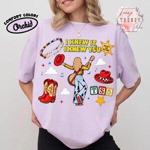 May include: A light purple t-shirt featuring a graphic print of a person with long hair playing a guitar, surrounded by cowboy boots, hats, a star, and blocks with the text I KNEW IT, I KNEW YOU, INFINITY AND BEYOND, and TS5.