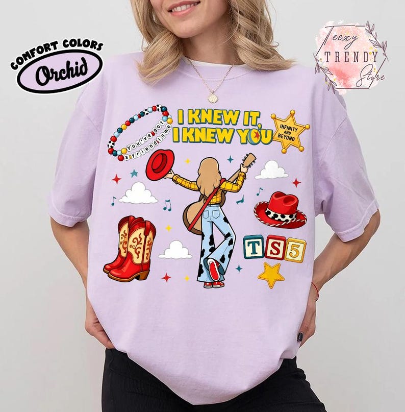 May include: A light purple t-shirt featuring a graphic print of a person with long hair playing a guitar, surrounded by cowboy boots, hats, a star, and blocks with the text I KNEW IT, I KNEW YOU, INFINITY AND BEYOND, and TS5.