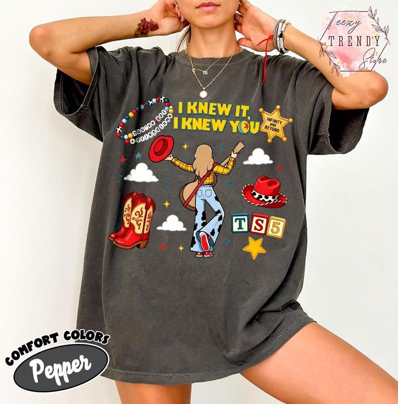 May include: A charcoal grey t-shirt featuring a graphic print with the text I KNEW IT, I KNEW YOU, INFINITY AND BEYOND, and TS5. The design includes a person with a guitar, red cowboy boots, a red hat, a beaded bracelet, and yellow stars.