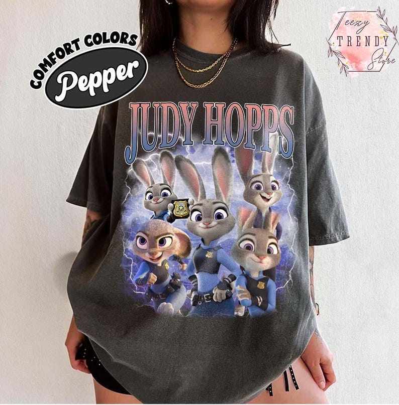 Puede incluir: Camiseta de color pimienta con un gr&aacute;fico de Judy Hopps de Zootopia. El dise&ntilde;o incluye m&uacute;ltiples im&aacute;genes de Judy Hopps con su uniforme de polic&iacute;a, con el texto "JUDY HOPPS" en degradado azul y rosa. La camiseta tiene un aspecto vintage y desgastado.