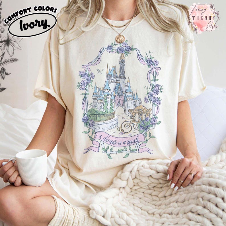 Puede incluir: Camiseta color marfil con un dise&ntilde;o de castillo en tonos pastel enmarcado por flores y lazos. El texto "a dream is a wish" est&aacute; en una pancarta. La camiseta tiene mangas cortas y un corte holgado.
