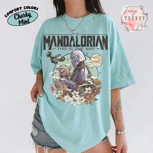 May include: A light blue t-shirt with a graphic from "The Mandalorian." The design includes the text "The Mandalorian This Is The Way" with an illustration of the Mandalorian, Grogu, and floral elements. The shirt has a relaxed fit.
