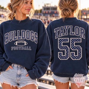 Op de afbeelding: Een marineblauw sweatshirt met ronde hals en een wit lettertype in universiteitsstijl met sterrenpatroon. De voorkant toont de tekst BULLDOGS FOOTBALL met een football-afbeelding. De achterkant toont de naam TAYLOR en het nummer 55.