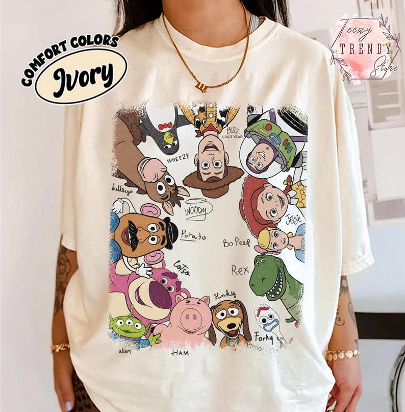May include: Ivory-colored t-shirt featuring a colorful graphic of various Toy Story characters, including Woody, Buzz Lightyear, and Jessie. The shirt has the words "COMFORT COLORS" and "Ivory" printed on it. The shirt is a crew neck.