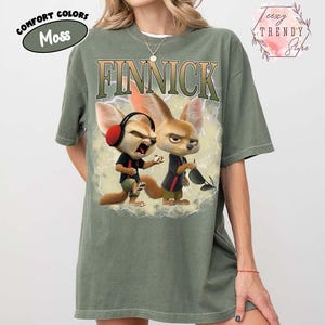 Puede incluir: Camiseta verde musgo con la palabra "FINNICK" en una fuente desgastada. La camiseta presenta un gr&aacute;fico de dos zorros de dibujos animados con auriculares y gafas de sol. La camiseta tambi&eacute;n tiene las palabras "Comfort Colors" y "Moss".