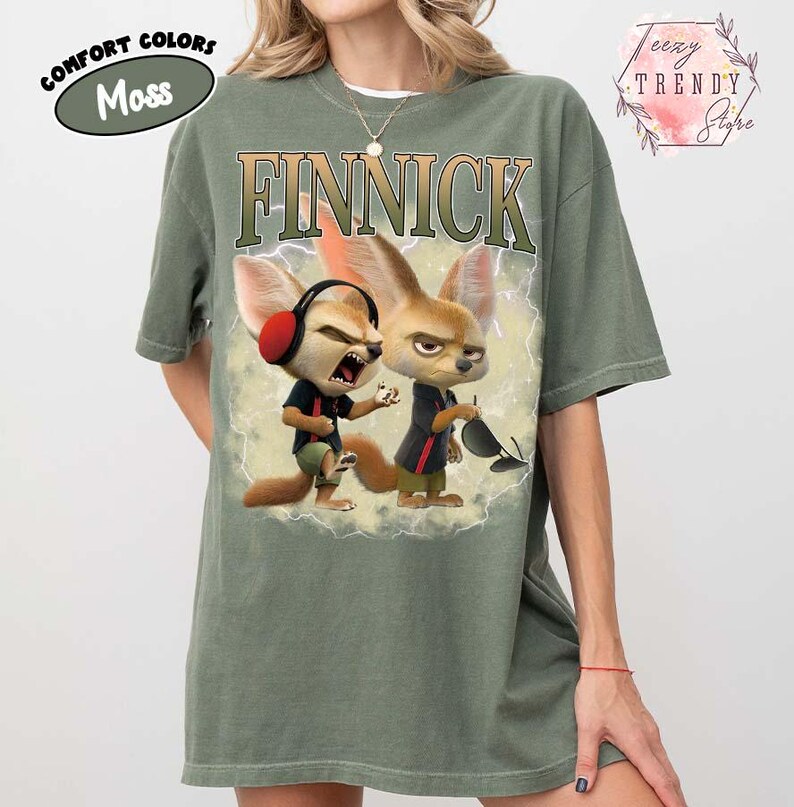 Puede incluir: Camiseta verde musgo con la palabra "FINNICK" en una fuente desgastada. La camiseta presenta un gr&aacute;fico de dos zorros de dibujos animados con auriculares y gafas de sol. La camiseta tambi&eacute;n tiene las palabras "Comfort Colors" y "Moss".