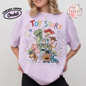 May include: A light purple t-shirt featuring a colorful graphic of characters from the animated film "Toy Story." The design includes the words "Toy Story" in a playful font, along with drawings and the Comfort Colors Orchid logo.