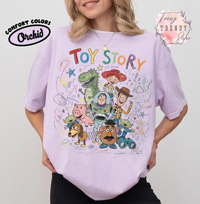 May include: A light purple t-shirt featuring a colorful graphic of characters from the animated film "Toy Story." The design includes the words "Toy Story" in a playful font, along with drawings and the Comfort Colors Orchid logo.