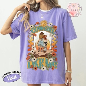May include: A violet short-sleeve t-shirt featuring a graphic print of two figures in cowboy hats and boots standing in a desert landscape. The design includes the text COWGIRL LIKE ME, surrounded by orange flames, white flowers, and a sun.