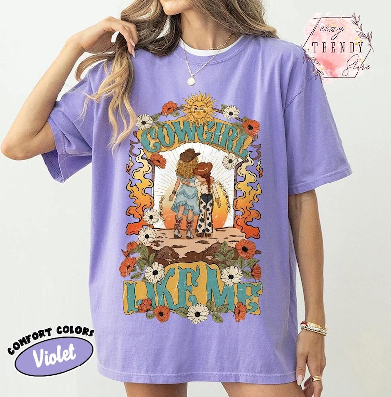 May include: A violet short-sleeve t-shirt featuring a graphic print of two figures in cowboy hats and boots standing in a desert landscape. The design includes the text COWGIRL LIKE ME, surrounded by orange flames, white flowers, and a sun.
