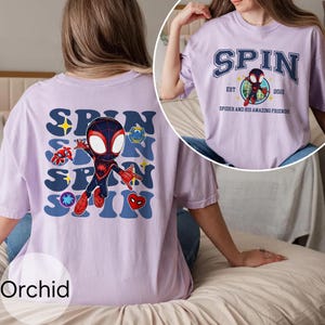 Puede incluir: Camisetas de color orqu&iacute;dea con un gr&aacute;fico de un Spider-Man de dibujos animados. La parte delantera de la camiseta tiene la palabra "SPIN" en una fuente estilizada, con un personaje de Spider-Man en el centro. La parte trasera de la camiseta tiene la palabra "SPIN" repetida con gr&aacute;ficos de Spider-Man.
