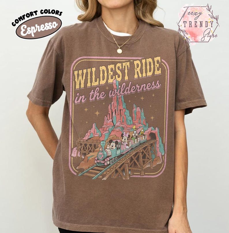 Może przedstawiać: T-shirt w kolorze espresso z grafiką w stylu vintage. Grafika głosi "Wildest Ride in the Wilderness" z pociągiem i postaciami. Koszulka ma okrągły dekolt i kr&oacute;tkie rękawy. Koszulka jest w kolorze komfortowym.
