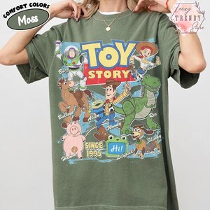 May include: A moss green short-sleeve t-shirt featuring a large graphic print of Toy Story characters including Woody, Buzz Lightyear, Jessie, Rex, Hamm, Bullseye, Slinky, and an Alien. The text reads Toy Story, Since 1995, and Hi on a lilypad.