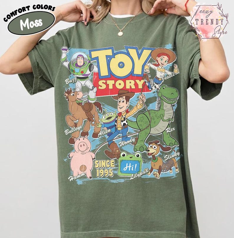 May include: A moss green short-sleeve t-shirt featuring a large graphic print of Toy Story characters including Woody, Buzz Lightyear, Jessie, Rex, Hamm, Bullseye, Slinky, and an Alien. The text reads Toy Story, Since 1995, and Hi on a lilypad.