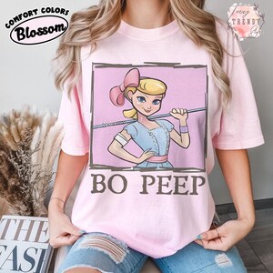 May include: A pale pink t-shirt with a cartoon image of Bo Peep from Toy Story. The design shows Bo Peep within a pink square frame, holding a shepherd's crook, with the words "BO PEEP" in brown. The shirt also has the words "Comfort Colors Blossom" printed on it.