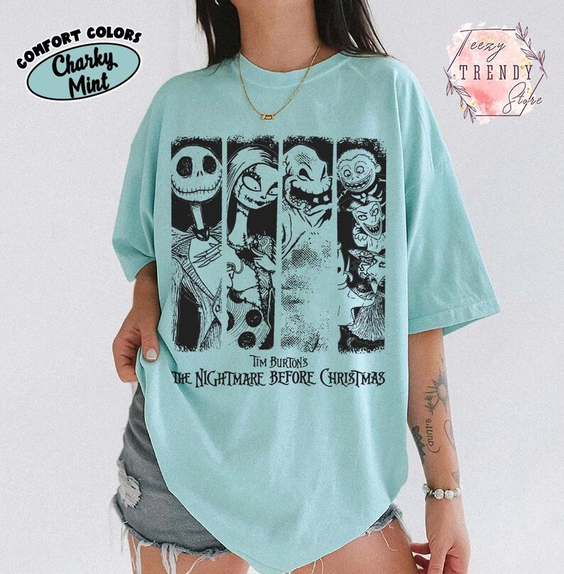May include: A light mint green short-sleeve t-shirt featuring a black graphic print of characters from Tim Burton's The Nightmare Before Christmas. The design includes four vertical panels showing Jack Skellington, Sally, Oogie Boogie, and Lock, Shock, and Barrel.