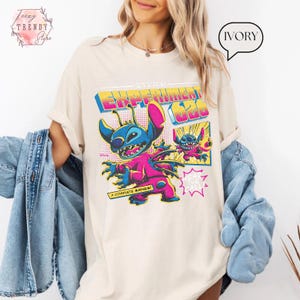Pode incluir: Camiseta de cor marfim com um gr&aacute;fico colorido de Stitch de Lilo & Stitch. O design inclui o texto "Stitch Experiment 626" e "A Complete Mayhem!" em um estilo retro de banda desenhada. A camiseta tem mangas curtas.