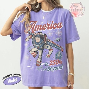 Puede incluir: Una camiseta violeta de manga corta con un gr&aacute;fico de Buzz Lightyear y el texto America To 250th and Beyond. El dise&ntilde;o incluye patrones de estrellas rojas, blancas y azules, fuegos artificiales y un planeta. La camiseta tiene cuello redondo y corte holgado.