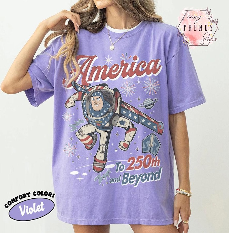 Puede incluir: Una camiseta violeta de manga corta con un gr&aacute;fico de Buzz Lightyear y el texto America To 250th and Beyond. El dise&ntilde;o incluye patrones de estrellas rojas, blancas y azules, fuegos artificiales y un planeta. La camiseta tiene cuello redondo y corte holgado.