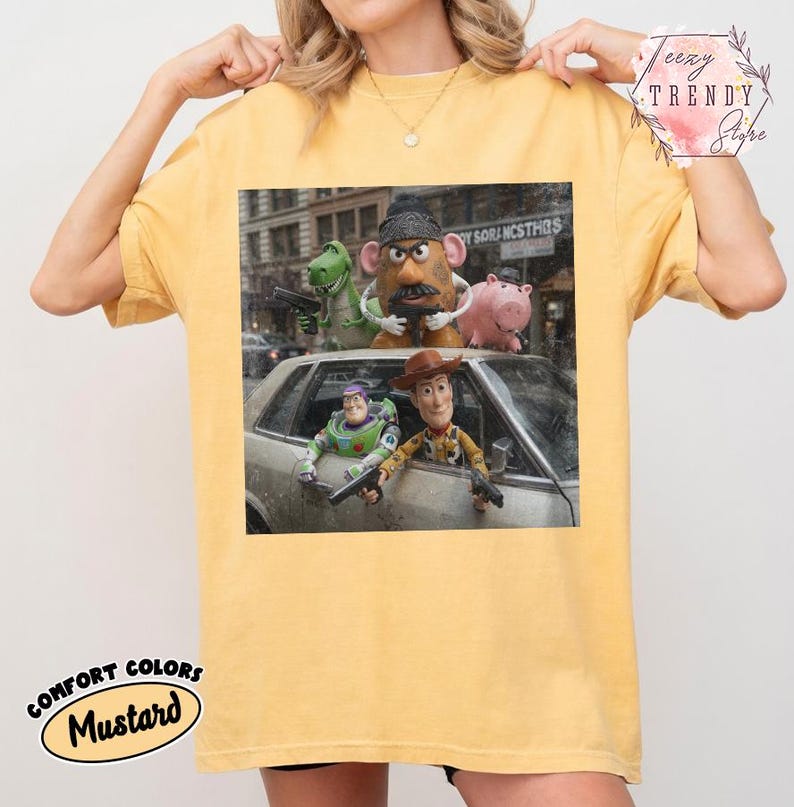 Puede incluir: Una camiseta de manga corta color amarillo mostaza con un estampado gr&aacute;fico de personajes de Toy Story, incluyendo a Woody, Buzz Lightyear, el Sr. Patata, Rex y Hamm, posando con pistolas de juguete frente a un coche en la ciudad.