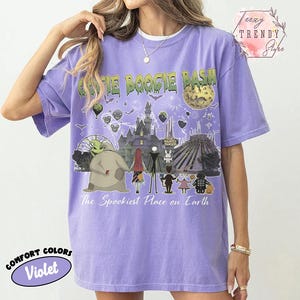 May include: A violet short-sleeve t-shirt featuring a graphic print with the text Oogie Boogie Bash and The Spookiest Place on Earth. The design includes illustrations of various characters, a castle, a moon, and fireworks in a dark theme.