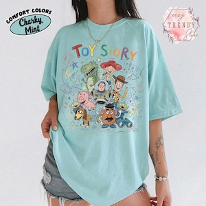 May include: A light blue t-shirt featuring the words "TOY STORY" above a colorful graphic of characters from the animated film. The shirt has a crew neck and short sleeves. The words "Comfort Colors" and "Charky Mint" are printed on the upper left.