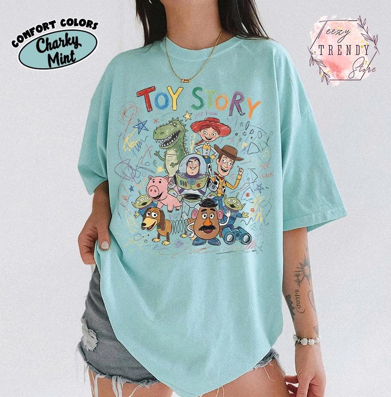 May include: A light blue t-shirt featuring the words "TOY STORY" above a colorful graphic of characters from the animated film. The shirt has a crew neck and short sleeves. The words "Comfort Colors" and "Charky Mint" are printed on the upper left.