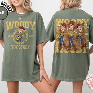 May include: Moss green t-shirt featuring Woody from Toy Story. The front design includes the word "WOODY" above a circular graphic of Woody, with "EST. 1995" and "TOY STORY" below. The back displays three Woody figures with the word "WOODY" repeated.