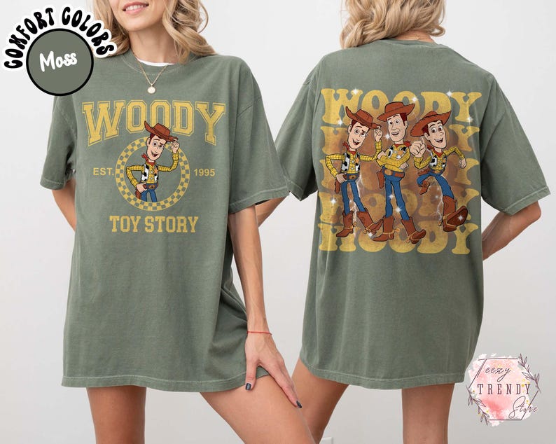May include: Moss green t-shirt featuring Woody from Toy Story. The front design includes the word "WOODY" above a circular graphic of Woody, with "EST. 1995" and "TOY STORY" below. The back displays three Woody figures with the word "WOODY" repeated.