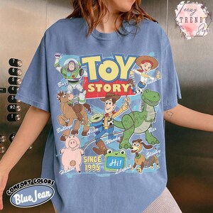 May include: A blue jean colored t-shirt featuring a graphic print of Toy Story characters including Buzz Lightyear, Jessie, Bullseye, Alien, Woody, Rex, Hamm, and Slinky Dog. The text reads Toy Story, Since 1995, and Hi! on a green lilypad.