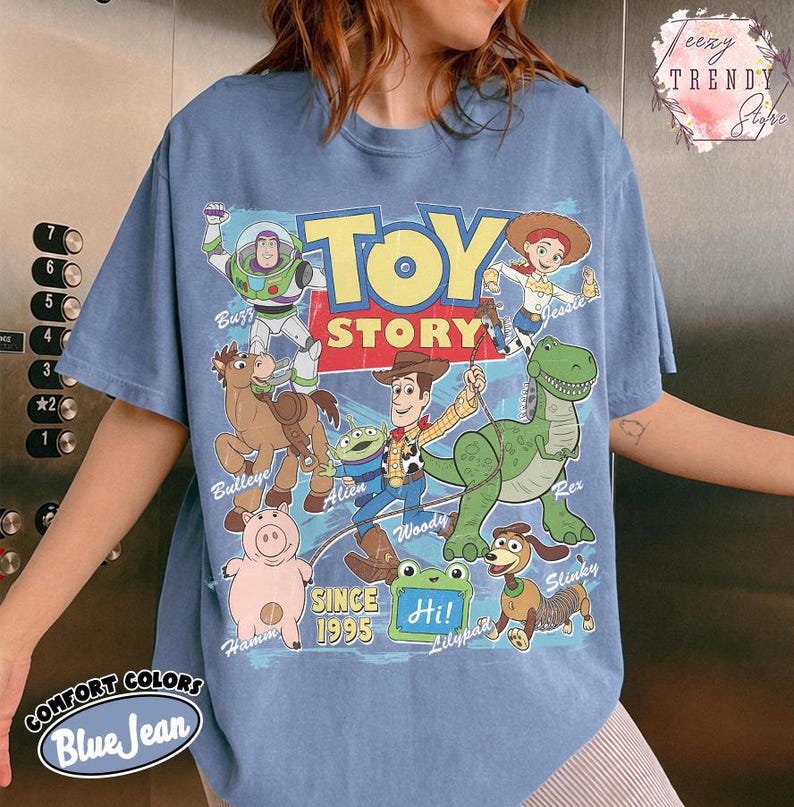May include: A blue jean colored t-shirt featuring a graphic print of Toy Story characters including Buzz Lightyear, Jessie, Bullseye, Alien, Woody, Rex, Hamm, and Slinky Dog. The text reads Toy Story, Since 1995, and Hi! on a green lilypad.
