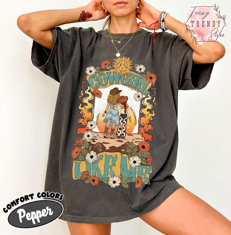 May include: A charcoal gray short-sleeve t-shirt featuring a graphic print of two figures in cowboy hats and boots standing in a desert landscape. The design includes orange flames, white flowers, a sun, and the text COWGIRL LIKE ME in teal letters.