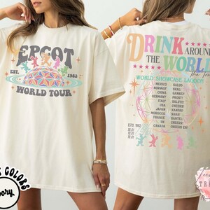 May include: Ivory-colored t-shirt featuring the text "EPCOT WORLD TOUR" with a colorful graphic of a geodesic sphere and characters. The back of the shirt reads "DRINK AROUND THE WORLD" with a list of countries and phrases.