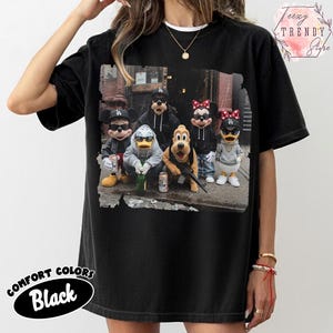 May include: A black short-sleeve t-shirt featuring a graphic print of cartoon characters including Mickey Mouse, Donald Duck, Goofy, Minnie Mouse, and Pluto wearing streetwear, sunglasses, and baseball caps while standing in an urban alleyway setting.