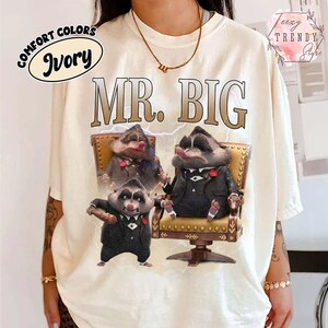 Puede incluir: Camiseta de color marfil con el texto "MR. BIG" en fuente marr&oacute;n. La camiseta presenta un gr&aacute;fico de tres mapaches con trajes, uno de ellos sosteniendo un cigarro. La camiseta tambi&eacute;n tiene las palabras "COMFORT COLORS" e "Ivory" en un c&iacute;rculo.
