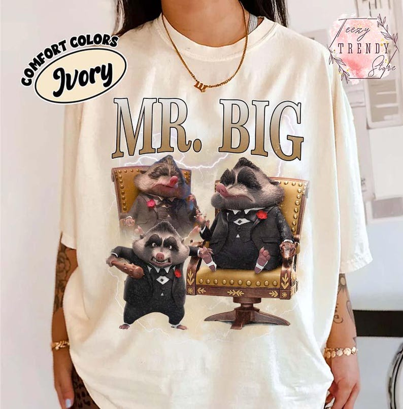 Puede incluir: Camiseta de color marfil con el texto "MR. BIG" en fuente marr&oacute;n. La camiseta presenta un gr&aacute;fico de tres mapaches con trajes, uno de ellos sosteniendo un cigarro. La camiseta tambi&eacute;n tiene las palabras "COMFORT COLORS" e "Ivory" en un c&iacute;rculo.