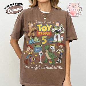 May include: An espresso-colored short-sleeve t-shirt featuring a graphic print of Disney Pixar Toy Story characters including Woody, Buzz Lightyear, Rex, and Forky. The text reads Toy Story 5 and You've Got a Friend In Me with a small Hi sign.