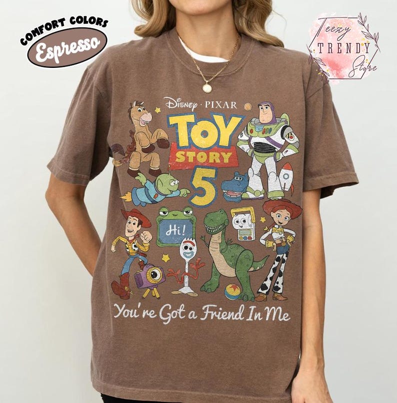 May include: An espresso-colored short-sleeve t-shirt featuring a graphic print of Disney Pixar Toy Story characters including Woody, Buzz Lightyear, Rex, and Forky. The text reads Toy Story 5 and You've Got a Friend In Me with a small Hi sign.