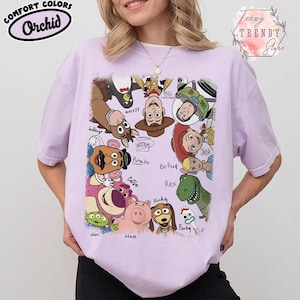 May include: Orchid Comfort Colors t-shirt featuring a colorful graphic of Toy Story characters. The design includes Woody, Buzz Lightyear, Jessie, and others. The shirt also has the text "eezy TRENDY Stighe".