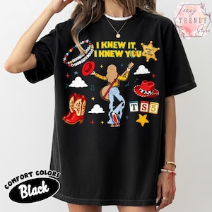 May include: A black t-shirt featuring a graphic print with the text I KNEW IT, I KNEW YOU, TS5, and INFINITY AND BEYOND. The design includes a central figure with a guitar, red cowboy boots, a red hat, a beaded bracelet, and colourful stars.